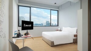 Habitación de hotel con cama, escritorio con silla, televisor y una ventana grande con vista a la ciudad.