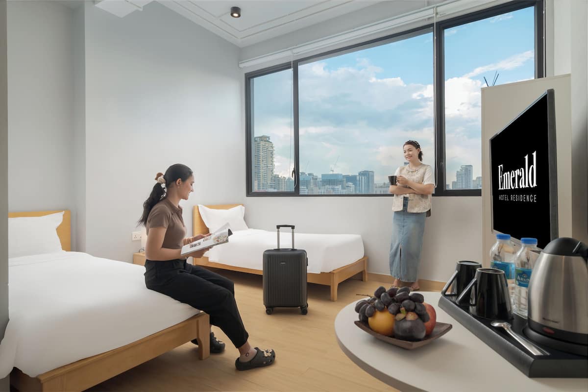 Ein Hotelzimmer mit Bett, Koffer, Obstschale und Fernseher, der "Emerald Hotel Residence" anzeigt.