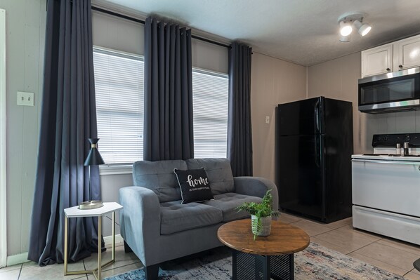 Una sala moderna con un sofá gris, un refrigerador negro, un horno blanco y una mesita redonda con una planta.