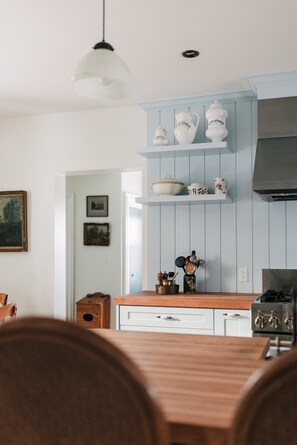 Une cuisine aux murs bleu clair, avec des étagères blanches où sont disposées des plats en céramique, une table à manger en bois et un luminaire suspendu.