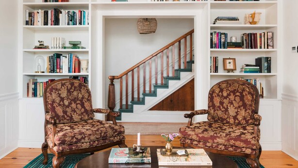 A living room with two armchairs, a coffee table, and a bookshelf filled with books. A staircase is visible in the background. - Little Louis - A Princely Take on a County Getaway (Prince Edward)