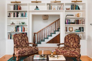 Un salon avec deux fauteuils, une table basse et une bibliothèque remplie de livres. On aperçoit un escalier au fond de la pièce.
