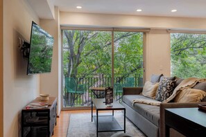 A living room with a sofa, a coffee table, a TV mounted on the wall, and a balcony with a view of trees. - Capitalia | Centric City Comfort -  Pet friendly (Ciudad de México)