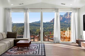 A living room with a sofa, coffee table, and a patterned rug. It features large sliding glass doors leading to an outdoor balcony with chairs and a table. - Epic views! Large uptown home w/ SwimSpa and Sauna (Sedona)