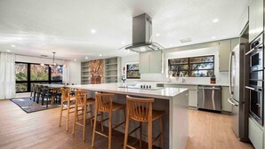 Una cocina moderna con una isla central, taburetes de bar, electrodomésticos de acero inoxidable y ventanas amplias.