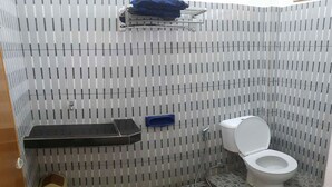 Une salle de bain avec un sol carrelé, des toilettes blanches, une poubelle bleue et une étagère en métal recouverte d’un tissu bleu.