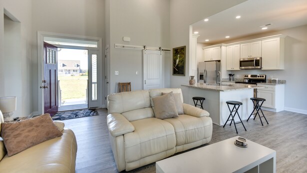 A modern living room with a beige leather sofa, a white coffee table, and a kitchen area with white cabinets and stainless steel appliances. - Cape Charles Home: 4 Mi to Kiptopeke State Park! (Cape Charles)