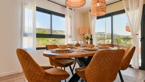 A dining area with a round table set for six, surrounded by six quilted chairs, large windows offering a view of greenery and hills, and two woven pendant lights. - Modern and comfortable apartment in Javea, Costa Blanca, Spain  with communal pool for 6 persons (Javea)