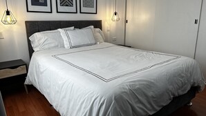 A neatly made bed with white bedding and black trim, a dark headboard, and a wooden floor. There are two wall-mounted light fixtures and three framed artworks above the bed. -  Aparts4Days 1 Bedroom Apartment San Isidro Olivar (Lima)