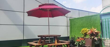 Un patio con mesa y bancos de madera, un paraguas rojo y plantas en macetas.