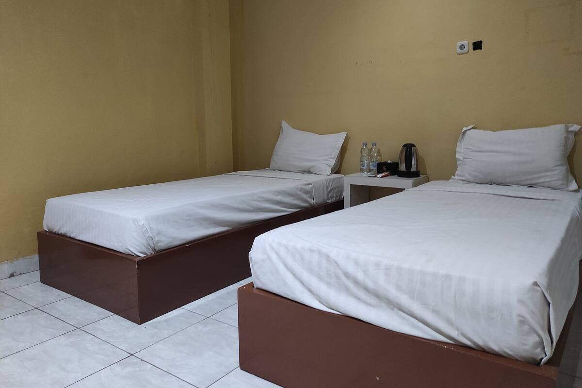 A hotel room with two single beds, a small table with a kettle, and a wall socket.