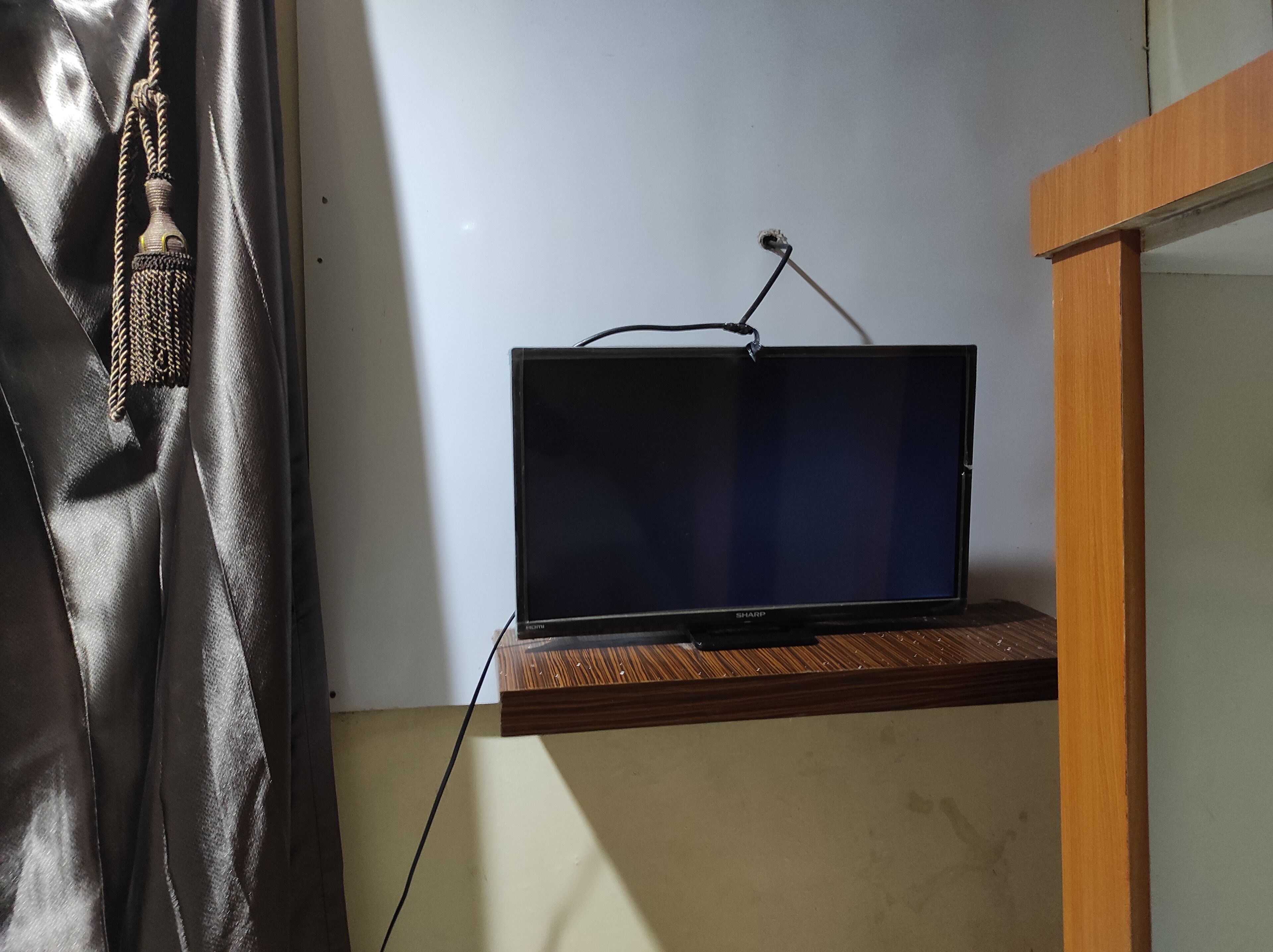 A television on a wooden shelf in a room with a plain wall and a black curtain.