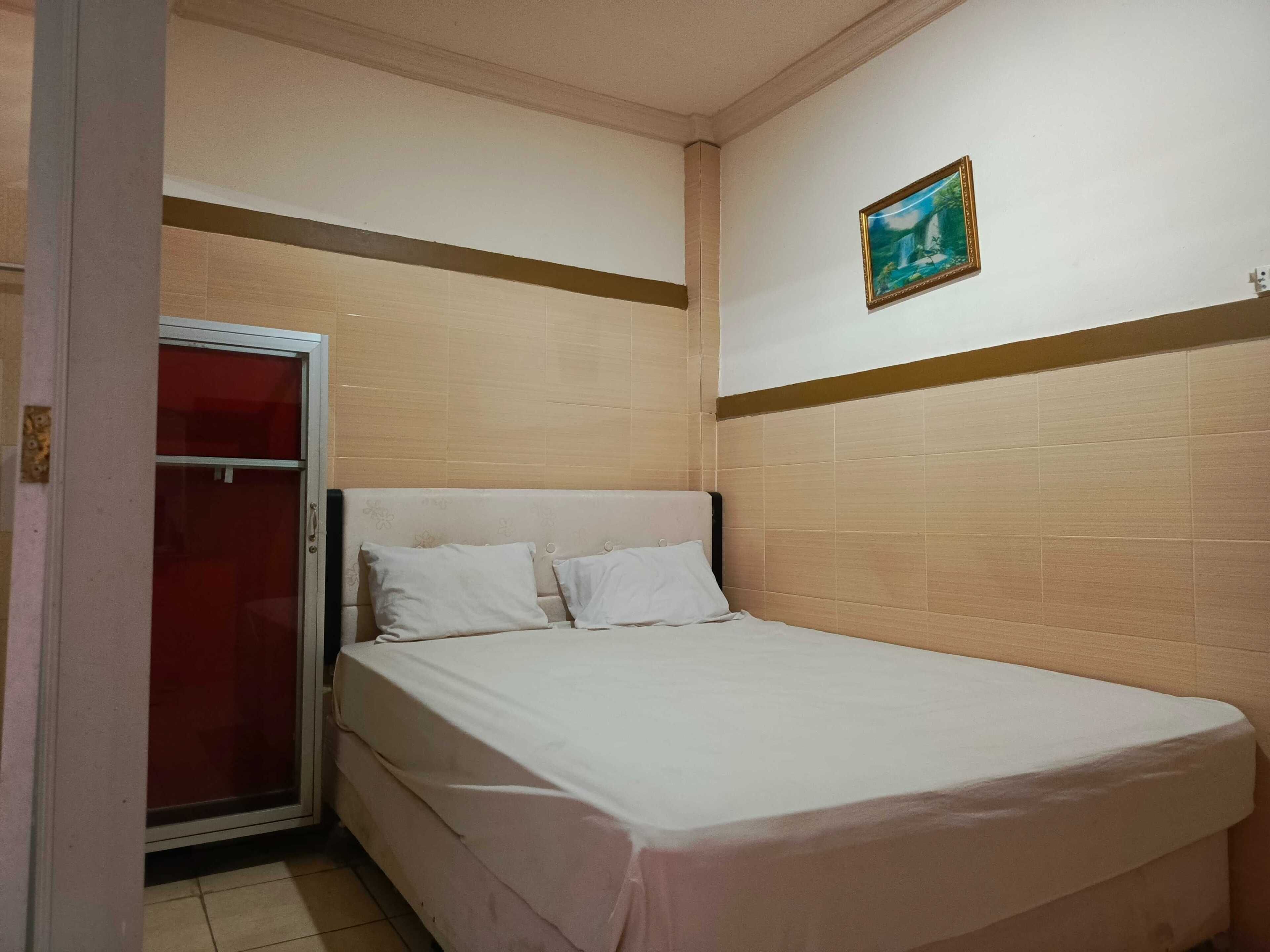 A small, single-bed room with a white bedspread, a wooden headboard, and a small painting on the wall.