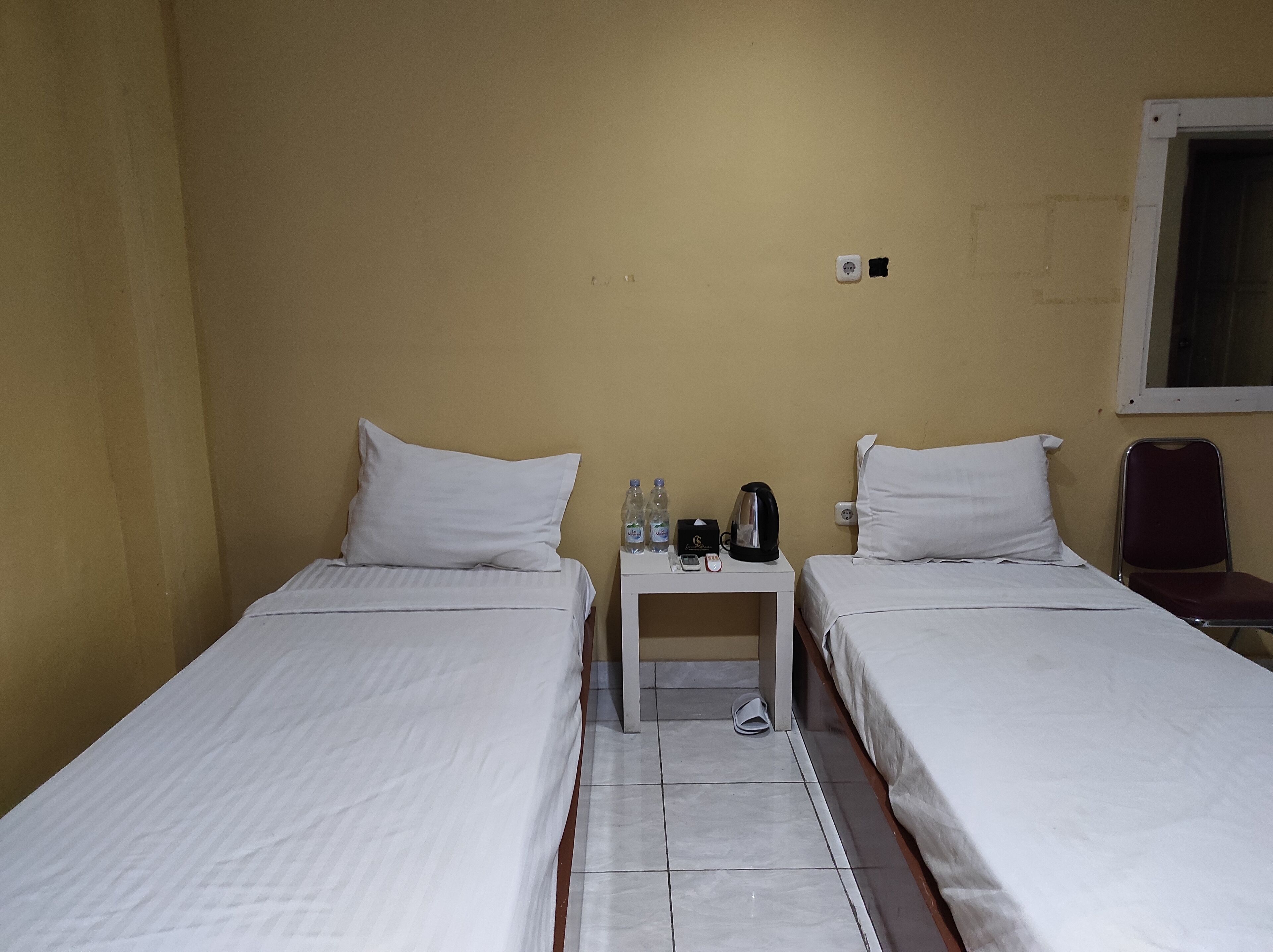 Quarto de hotel com duas camas de solteiro, uma mesinha com chaleira, telefone e garrafas de água.