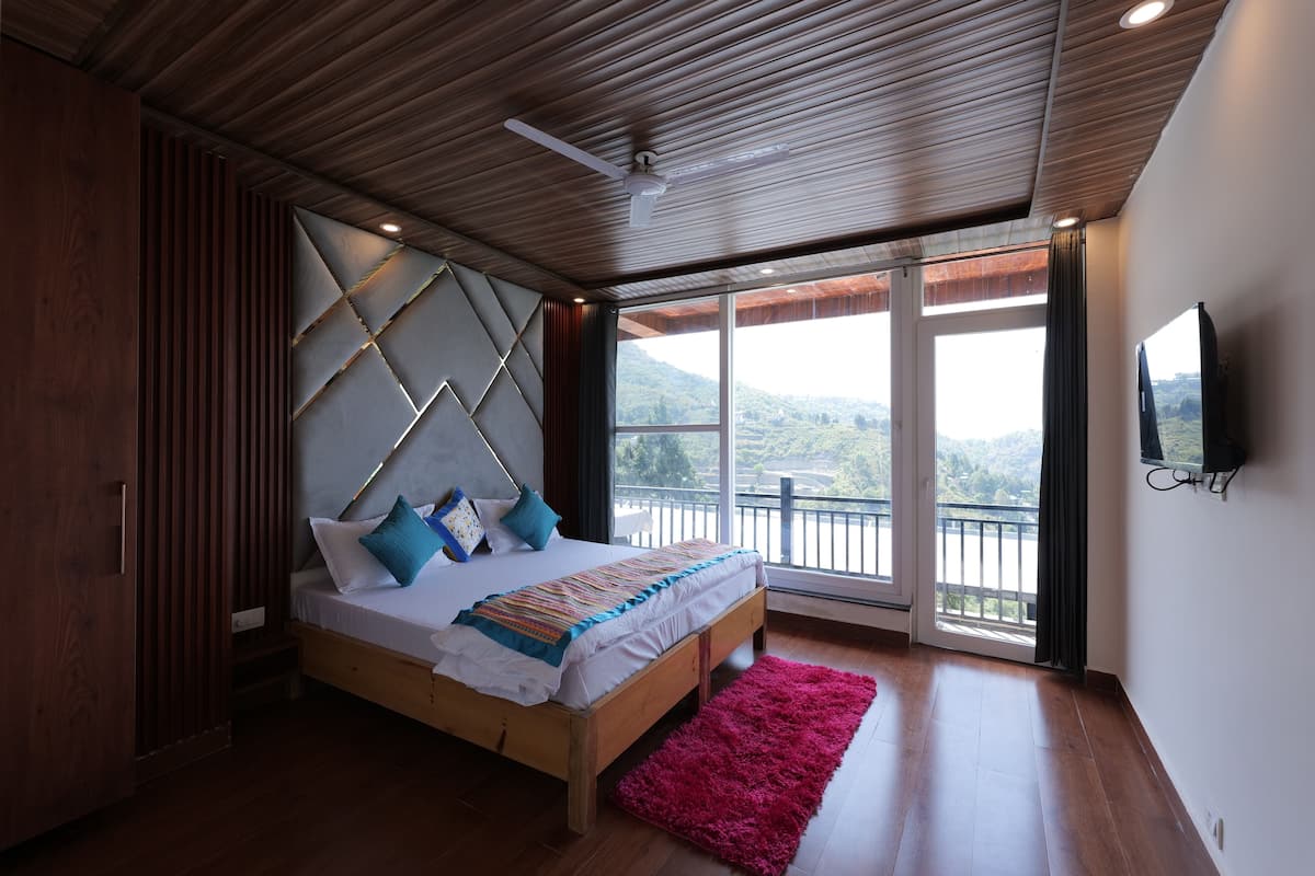 Un dormitorio con techo de madera, una cama con colcha estampada, un tapete rosa y un ventanal con vistas al exterior.