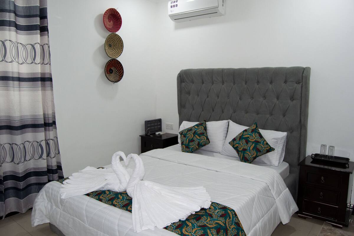 A neatly made bed with white linens and a decorative towel arrangement, a grey tufted headboard, and a bedside table with a radio.