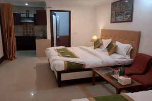 A hotel room with a bed, a small table with a bottle and a box, a red chair, and a view of a kitchen and bathroom through an open door. - Hotel Shyam Palace (New Delhi)