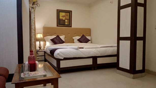 A bedroom with a bed, bedside table, lamp, and a painting on the wall. - Hotel Shyam Palace (New Delhi)