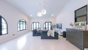Un salon spacieux avec un grand canapé, une commode et un téléviseur. De grandes fenêtres et un luminaire au plafond complètent l’ensemble.