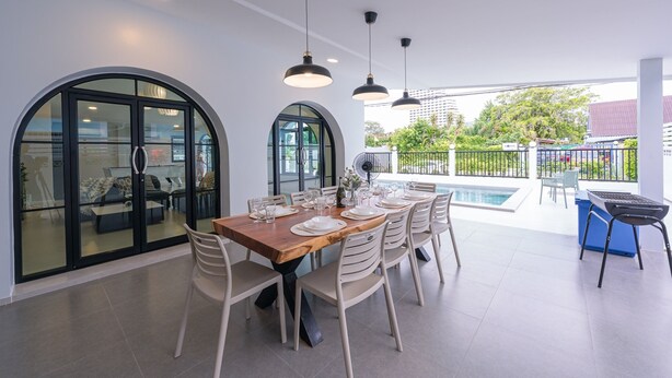 Un coin repas avec une table en bois dressée pour six personnes, entourée de chaises blanches, et en arrière-plan, on aperçoit une piscine et un espace salon extérieur.