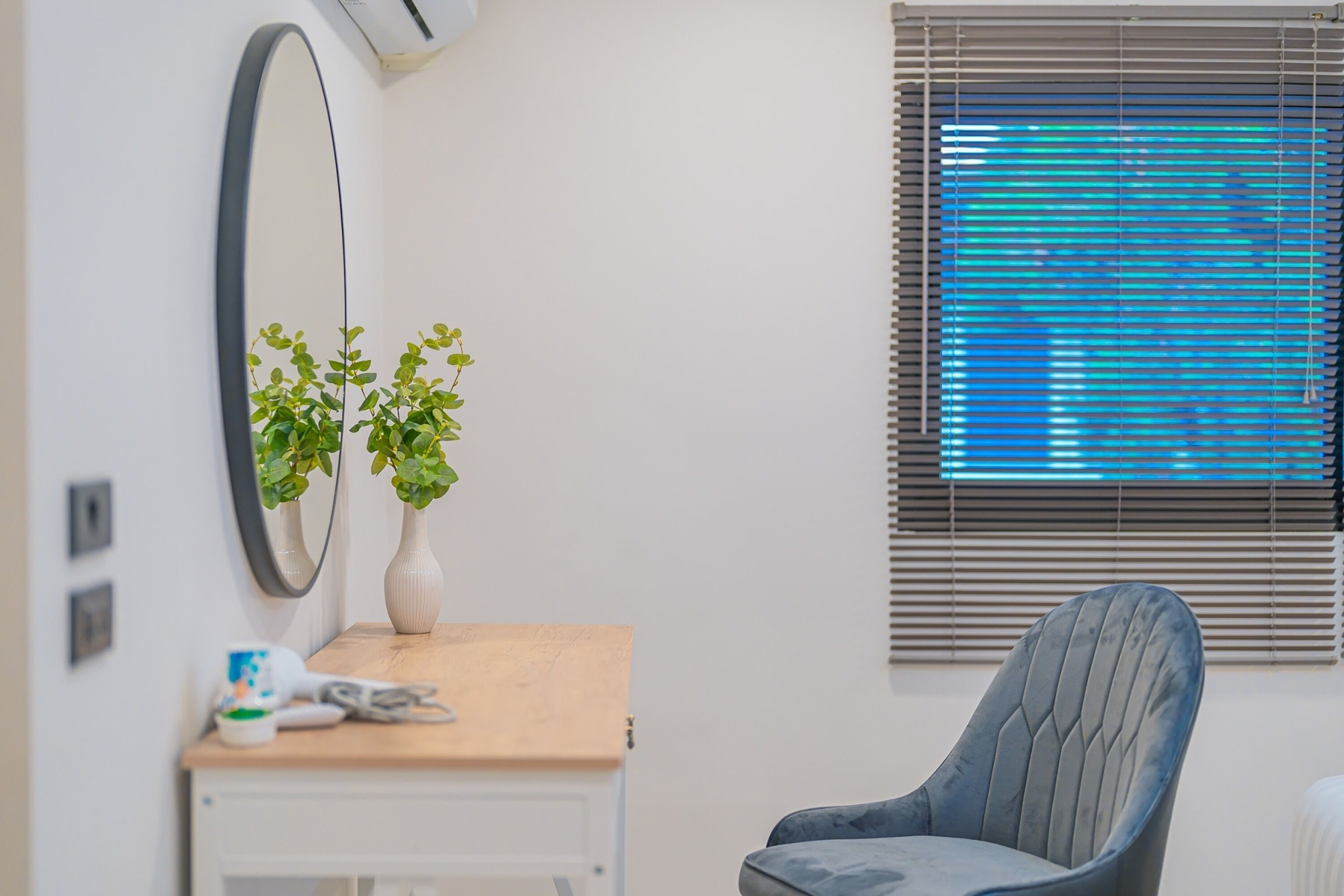 Une pièce comprenant un bureau en bois, une chaise, un miroir, deux plantes en pot et une fenêtre avec des stores.