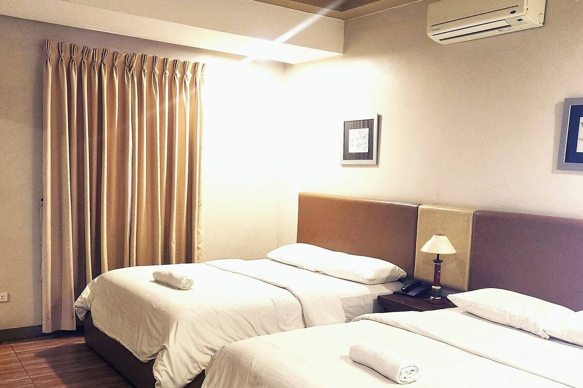 A hotel room with two beds, a ceiling fan, an air conditioning unit, a bedside lamp, and a window with curtains.