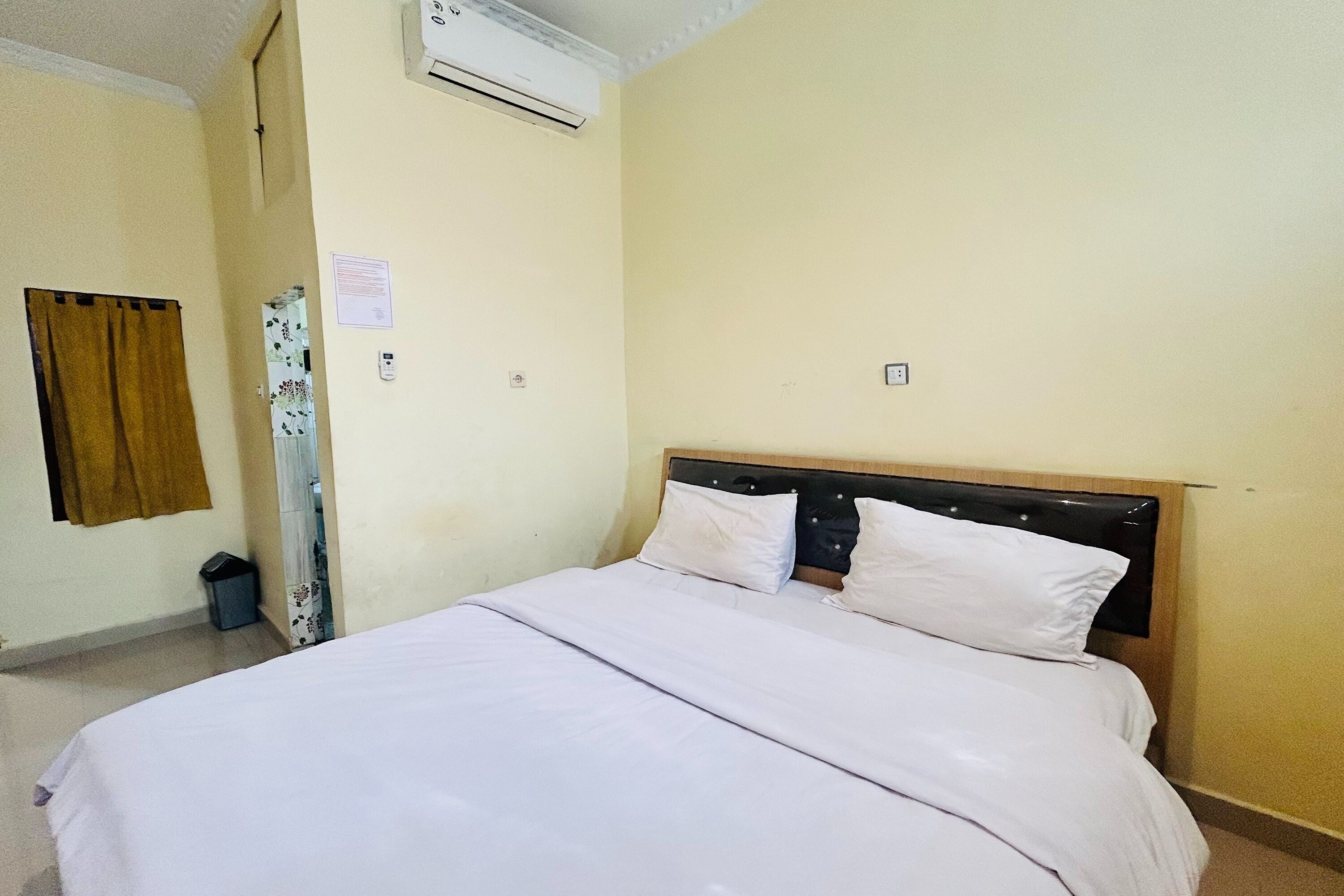 A hotel room with a bed, a wall-mounted air conditioner, a trash bin, and a curtain.