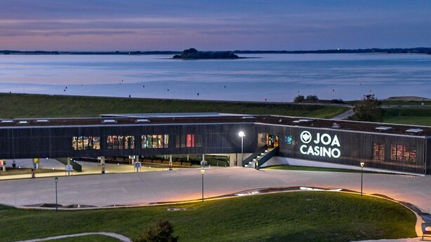 A modern building with a prominent "JOA CASINO" sign, surrounded by a well-maintained grassy area and a body of water in the background.