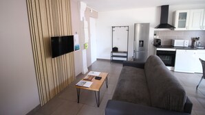 A modern living room with a flat-screen TV, a wooden accent wall, a sofa, a coffee table, and a kitchen area with appliances.