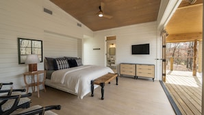 Ein Schlafzimmer mit Bett, Nachttisch, Lampe und einer Bank. Außerdem ein Schminktisch und ein an der Wand befestigtes Fernsehgerät.