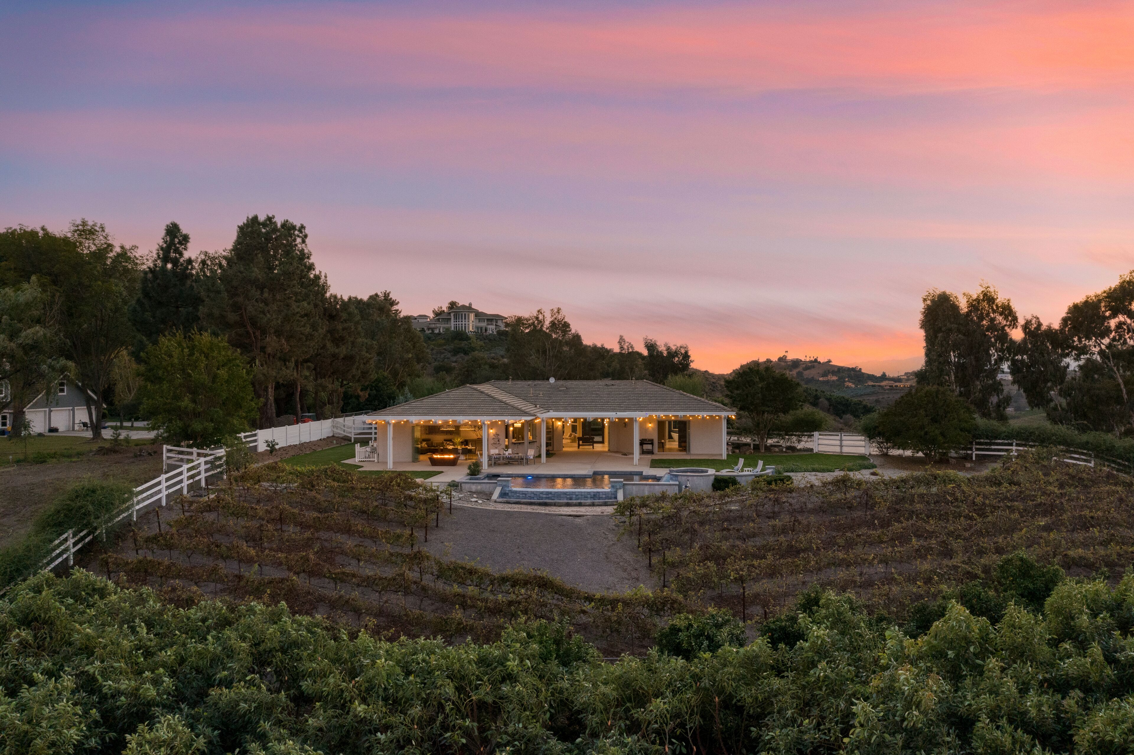 A large, modern house with a spacious patio and a swimming pool, surrounded by lush greenery and hills.