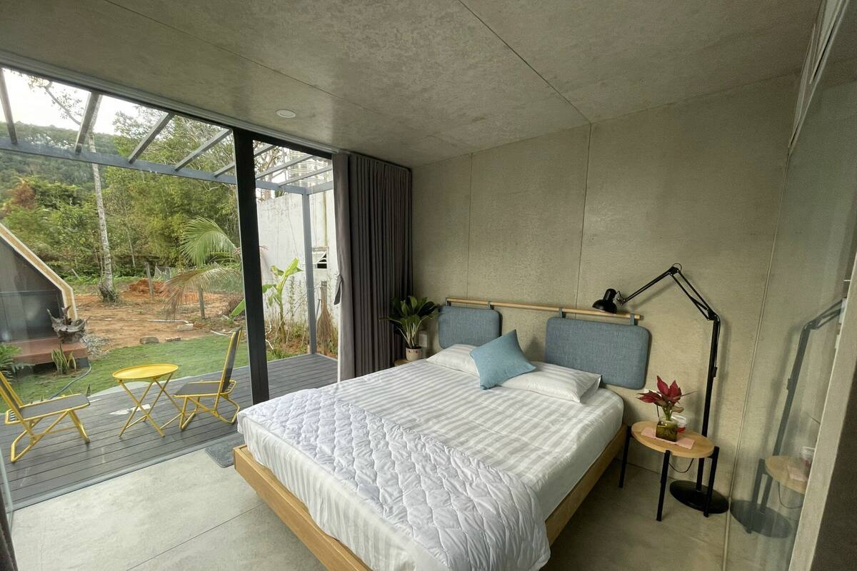 A modern bedroom with a large bed, a bedside table with a lamp, and a view of a garden through a sliding glass door.