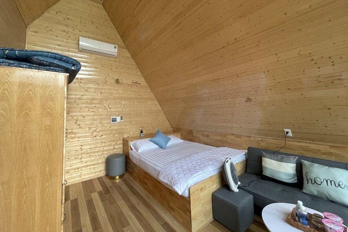 A cozy, wooden-paneled room with a bed, a sofa, a small table, and a wall-mounted air conditioner.