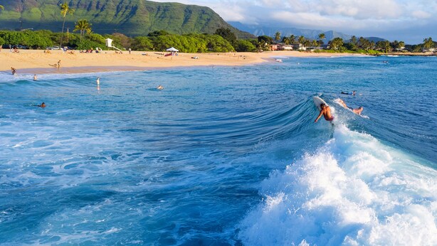 A beach with surfers, clear blue water, and a mountainous backdrop.
