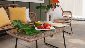 A glass-top table with a fruit platter, wicker chairs, and a potted plant on a patio.