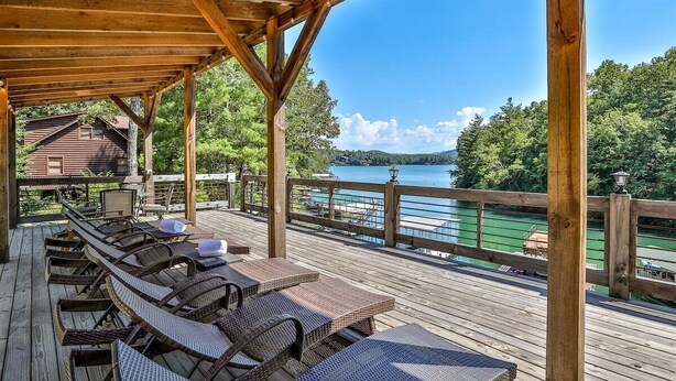 A covered outdoor deck with lounge chairs and tables, overlooking a lake and forested area. - Live Love Lake | Blue Ridge, GA (Blue Ridge)