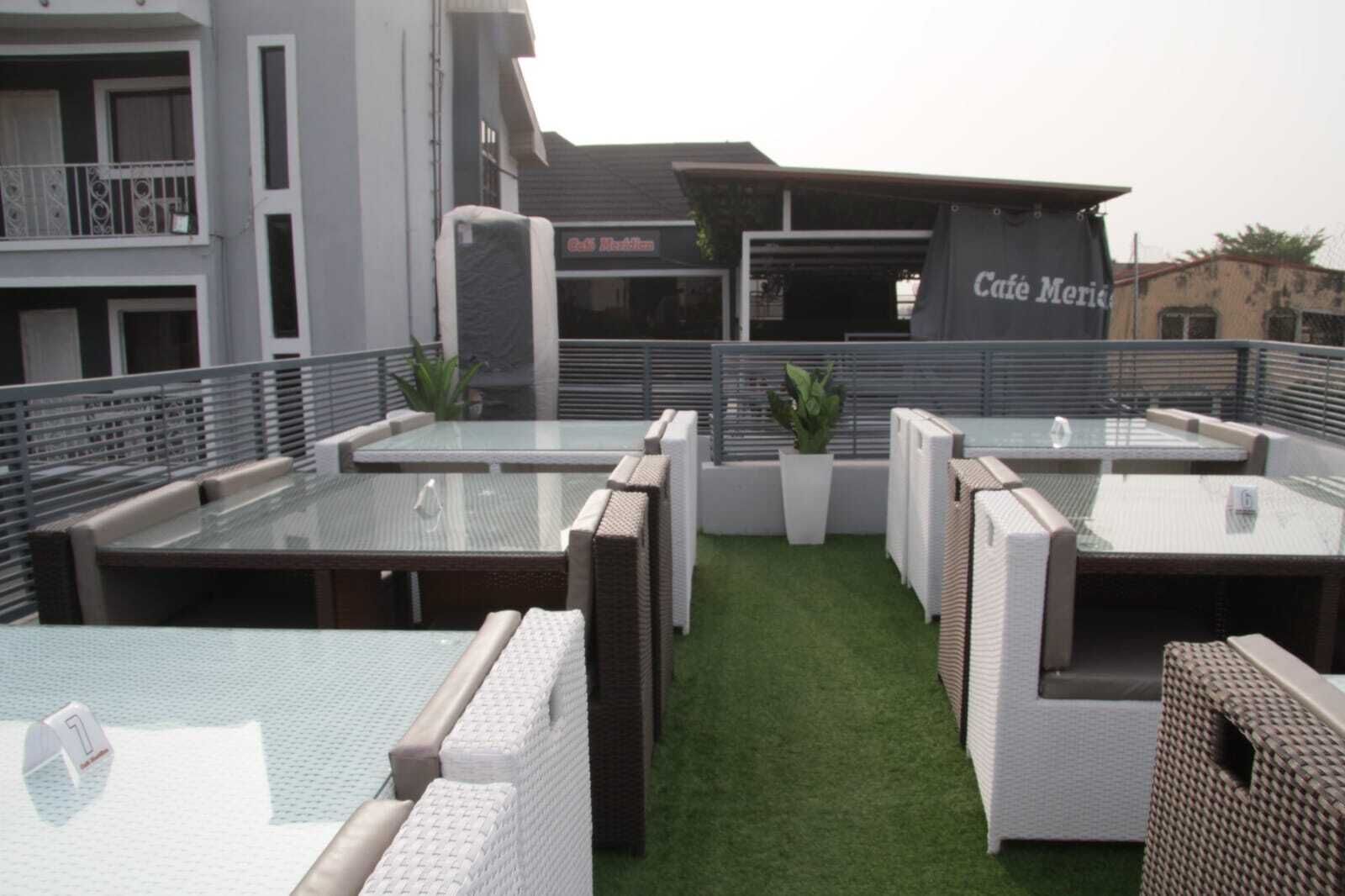 A rooftop terrace with glass-top tables and wicker chairs, a sign for 'Cafe Merida', and a building with balconies.