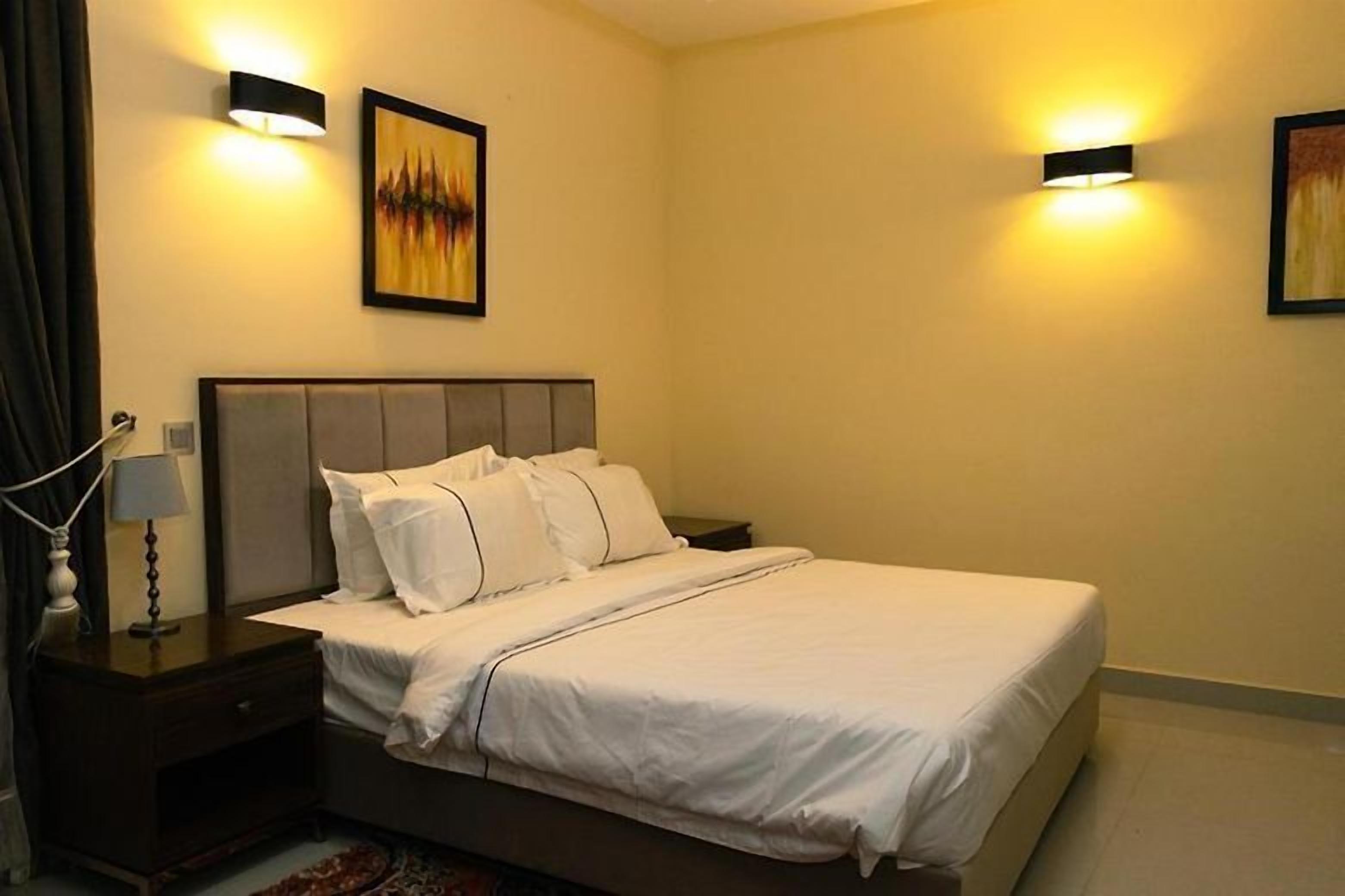 A neatly made bed in a hotel room with a headboard, two wall-mounted lamps, and a framed picture on the wall.