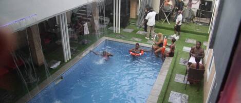 A swimming pool with people around it, a covered area with a stage, and a building with a balcony.