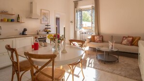 A modern kitchen with white cabinets, a dining area with a round table and chairs, a sofa, and a large window with curtains. - Vacation home Randi by Interhome (Anzio)