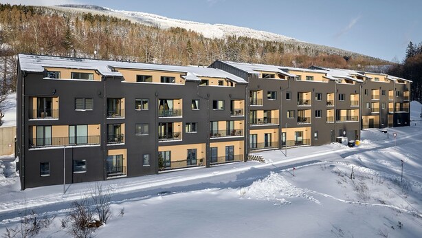 Un moderno edificio de apartamentos de varios pisos, con balcones, situado en una zona nevada y con una montaña al fondo.