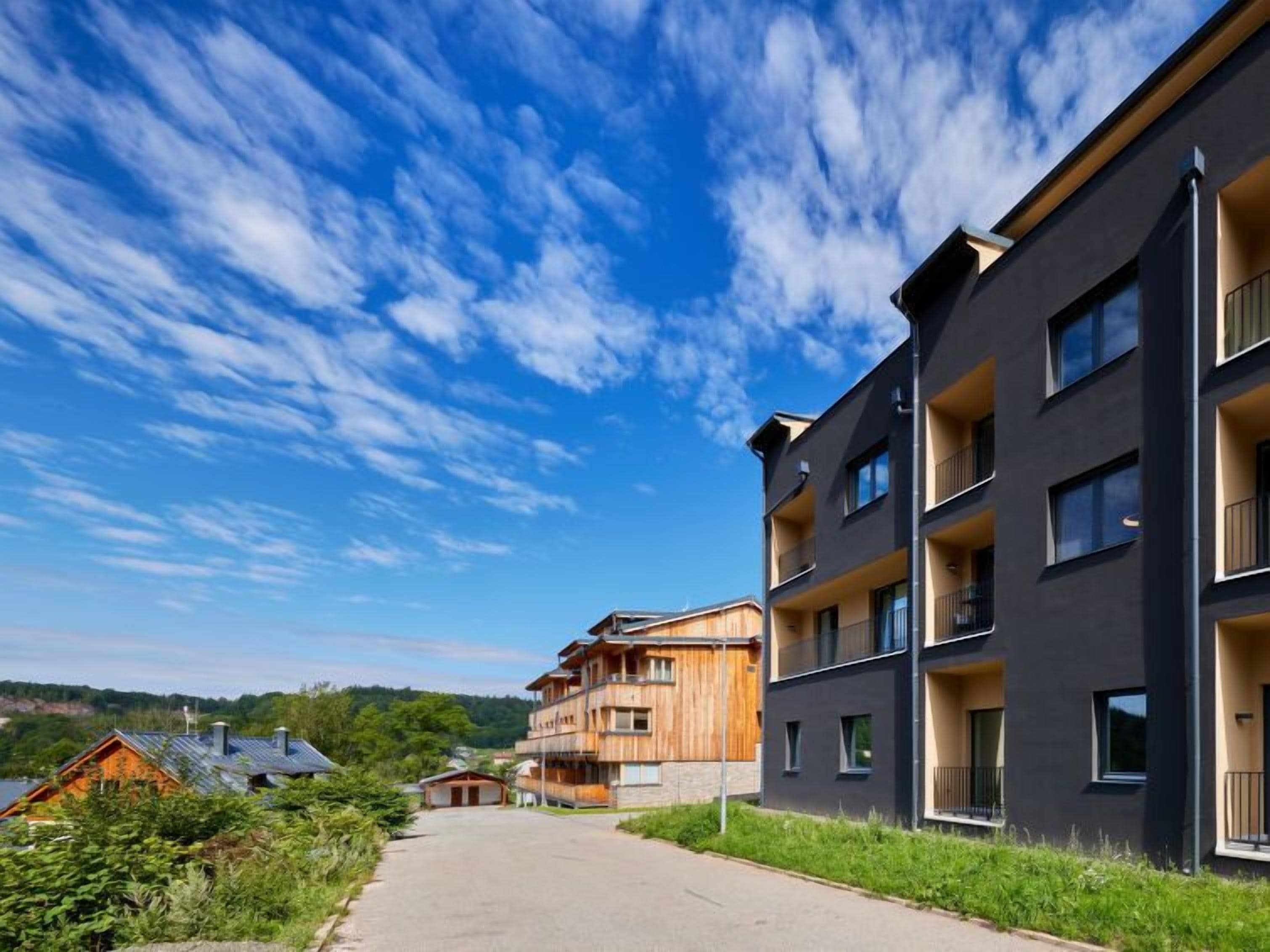 Um edifício de apartamentos moderno com várias varandas, céu claro e um caminho que leva a um prédio de madeira ao fundo.