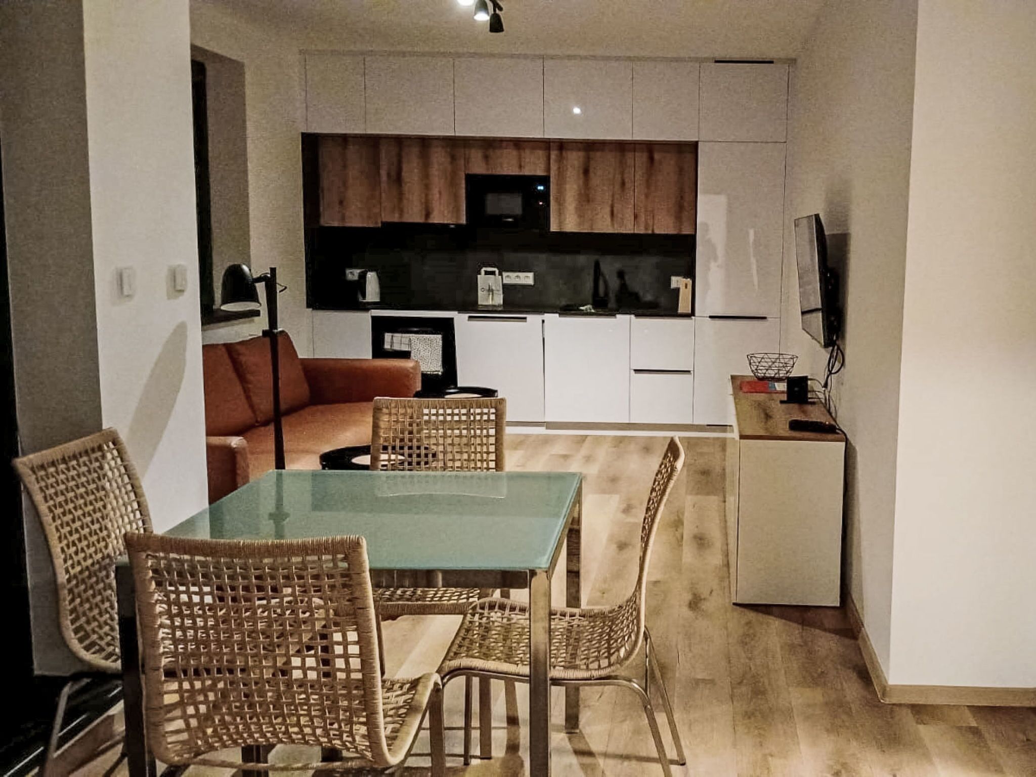 Ett kompakt bostadsutrymme med ett matbord, flätade stolar, en soffa, ett litet kök och en tv.