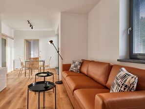 Een moderne woonkamer met een leren bank, twee zwarte bijzettafels, een eethoek met een tafel en stoelen, en een vloerlamp.
