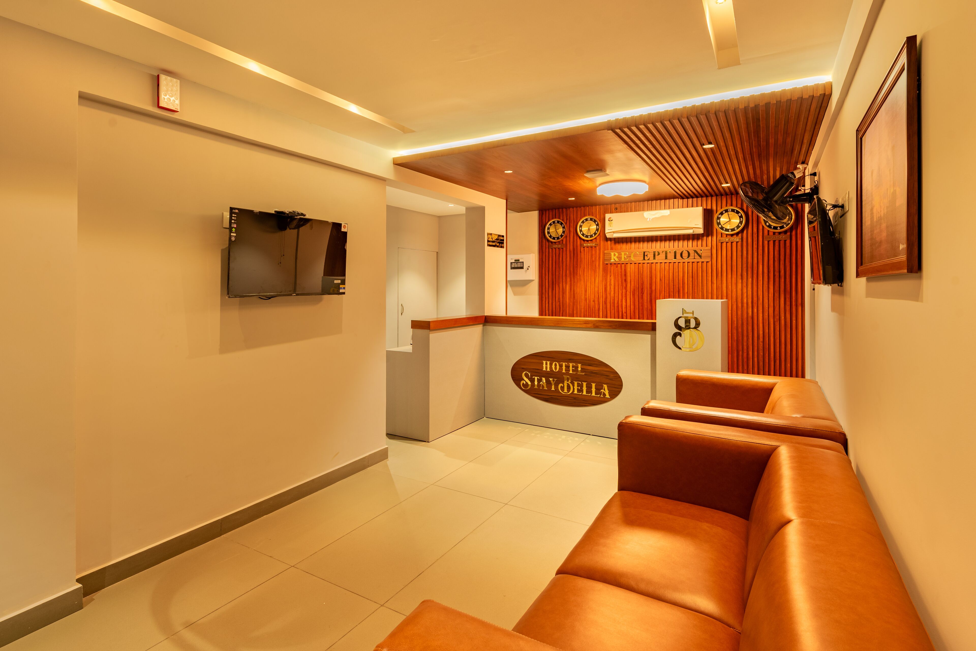 A hotel reception area with a wooden reception desk, a television mounted on the wall, and a seating area with leather sofas.