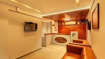 A hotel reception area with a wooden reception desk, a television mounted on the wall, and a seating area with leather sofas.