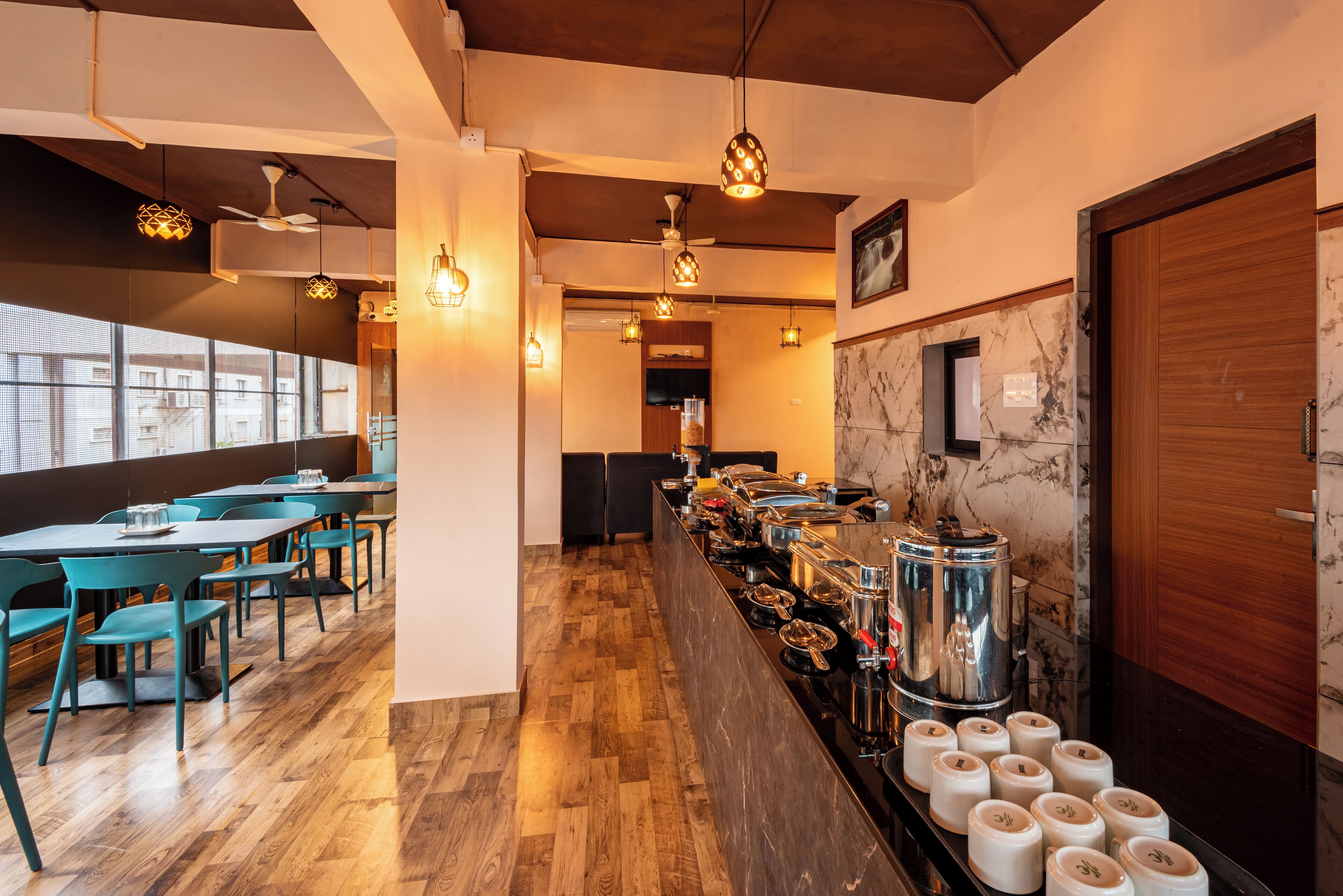 Un restaurante con suelo de madera, sillas color turquesa, un mostrador de buffet con varios recipientes de comida y lámparas colgantes.