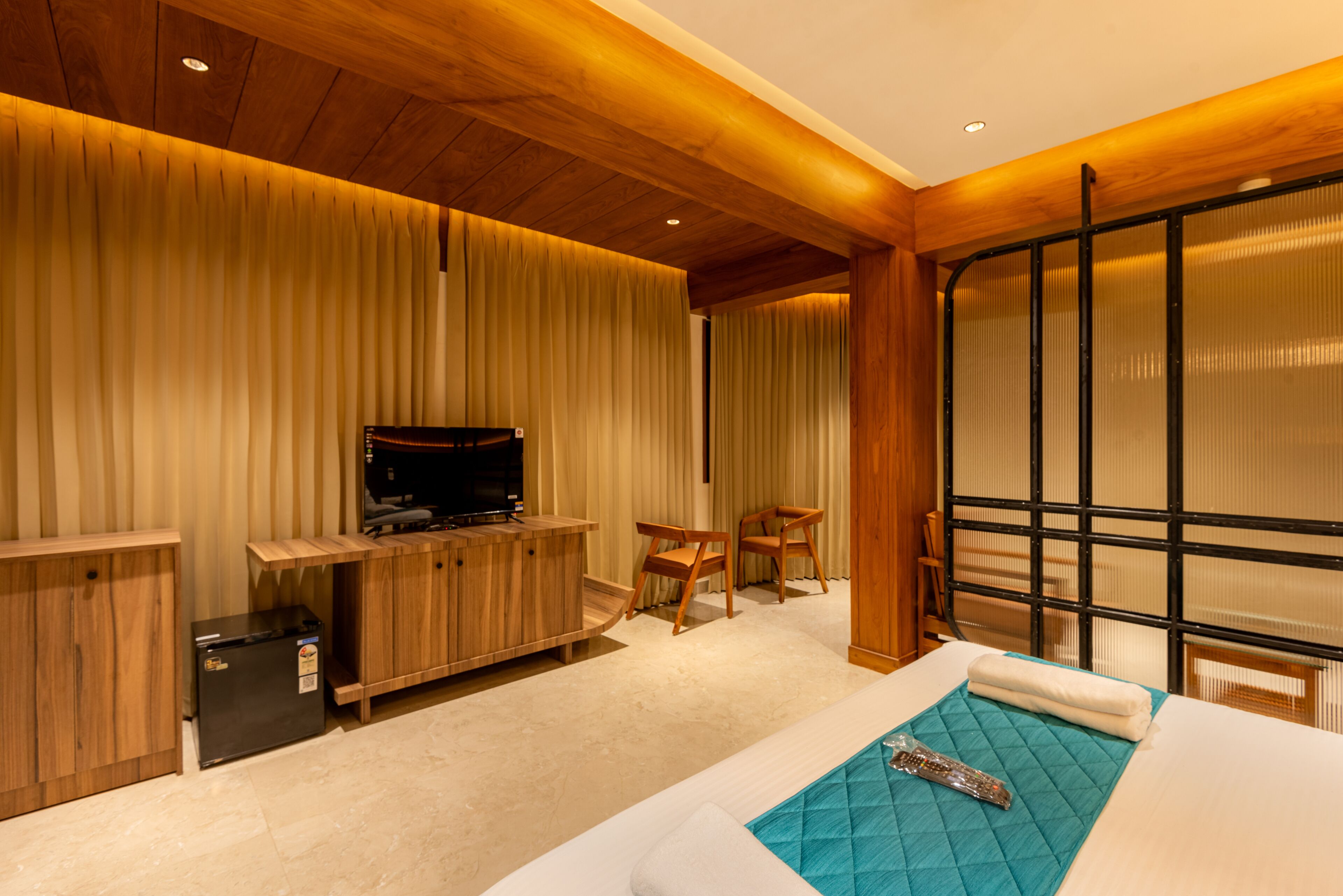 A modern hotel room with a wooden ceiling, a flat-screen TV on a wooden cabinet, a small refrigerator, a bed with a blue quilt, and a large window with blinds.
