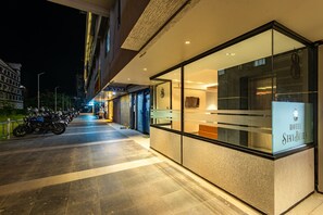 A hotel entrance with a glass facade, a row of parked motorcycles, and a visible ATM sign. - Hotel Stay Bella (Kozhikode)