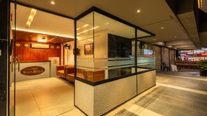 Zona de recepción de hotel con una pared de vidrio, mostrador de recepción y área de espera con sillones de cuero.